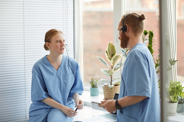 Two medical workers in uniform and headphones standing at office and discussing work together