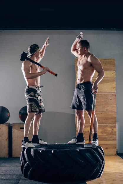 Two men doing high five while standing on tire and holding asledgehammer