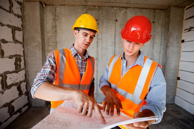 Two men dressed in orange work vests and helmets explores construction documentation on a concrete wall background .