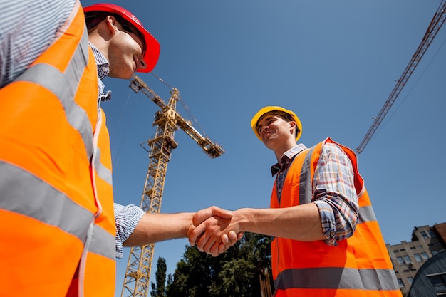 Two men dressed in orange work vests and helmets shake hands on the building site near the crane 