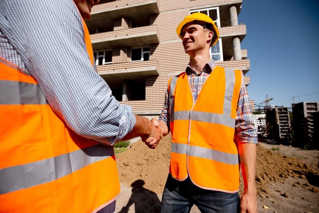 Two men dressed in shirts, orange work vests and helmets shake hands against the background of a multistorey building .