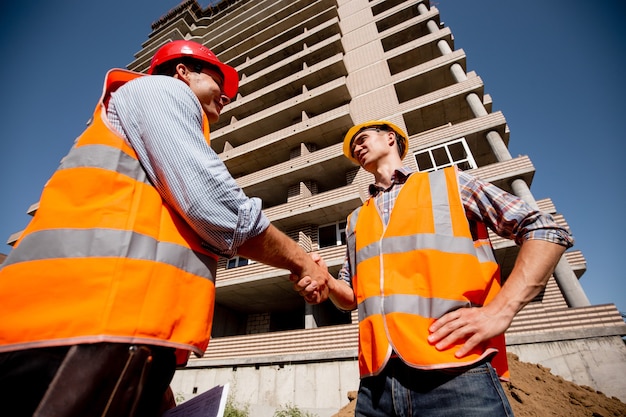 Photo two men dressed in shirts, orange work vests and helmets shake hands against the background of a multistorey building .