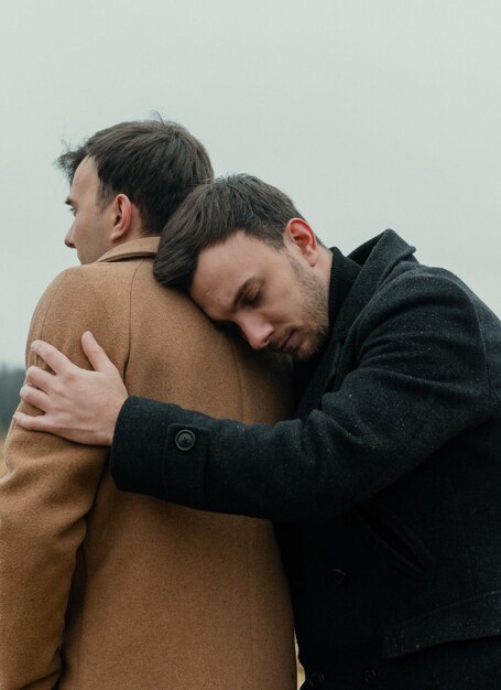 Two men hugging each other, one of which is wearing a black coat and the other wearing a brown coat.