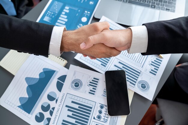 Two men shaking hands over a desk with charts and graphs on the table.