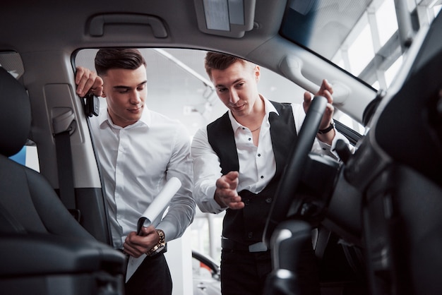 Two men stand in the showroom against cars. Close-up of a sales manager in a suit that sells a car to a customer. The seller gives the key to the customer.