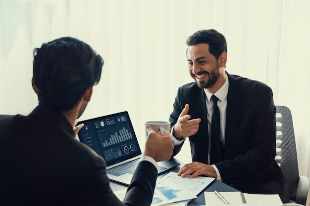 Two men in suits are sitting at a desk, one of them is giving a thumbs up to a laptop with a graph showing the time of the year.