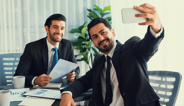 Two men in suits are sitting at a desk, one of them is taking a self portrait.