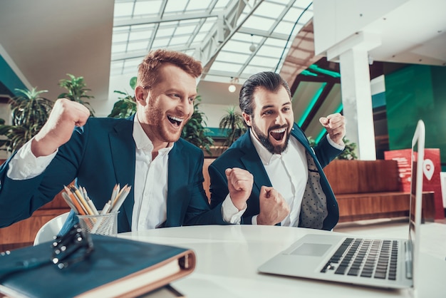 Two men in suits celebrating looking on laptop in office.