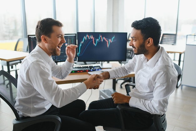 Two men traders sitting at desk at office together monitoring stocks data candle charts on screen analyzing price flow smiling cheerful having profit teamwork concept