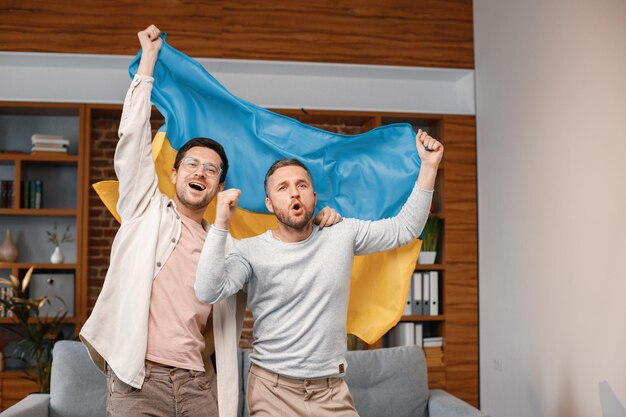 Two men watching a football game on tv and holding big flag of Ukraine