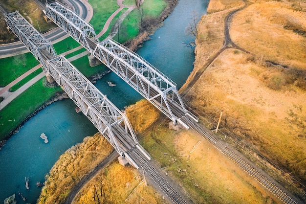 Two metal bridges, with a railway web, over a small river, one\
bank green, with lawn, the other yellow, with dry grass.\
contrasting landscape. drone view.