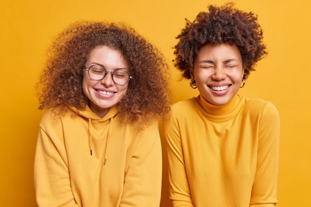 Two mixed race women with curly hair have happy expressions giggle positively stand next to each other close eyes from joy spend free time together isolated over yellow wall. Emotions concept