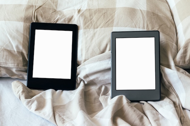 Two modern electronic books with a blank empty screens on a white and beige bed. tablets on bedding