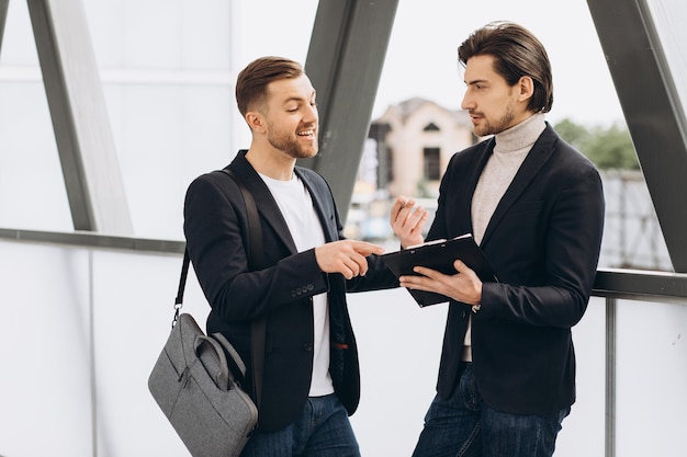 Photo two modern happy businessmen with documents folder discussing something against the background of urban offices and buildings