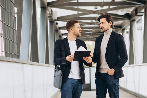 Two modern happy businessmen with documents folder discussing something against the background of urban offices and buildings