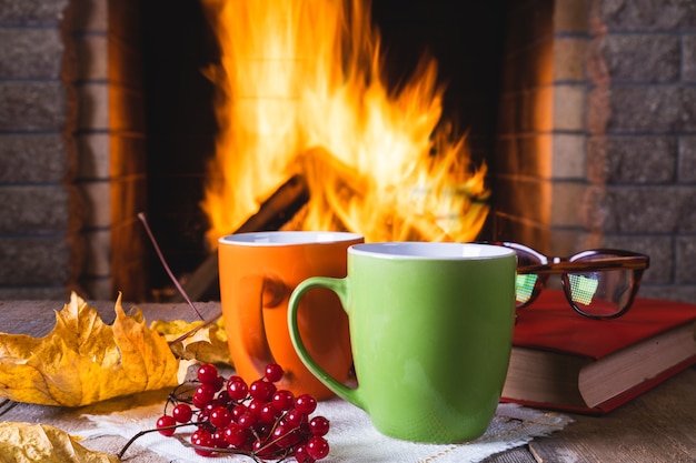 Two mugs of tea or coffee, autumn leaves and viburnum berries near a cozy fireplace, in a country house, autumn or winter holidays,horizontal.