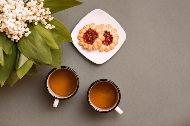 お茶と2つのマグカップ、皿にクッキー、緑のテーブルにスズランの花束。休息のために一時停止し、人生を遅くします。上面図