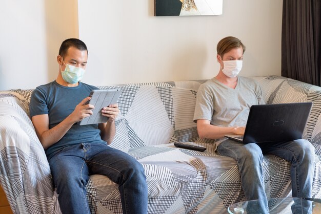 Two multi ethnic men with mask as friends using technology in quarantine at home