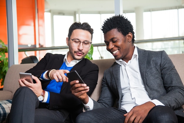 Two multicultural men sit on sofa, smile and hold mobile phones