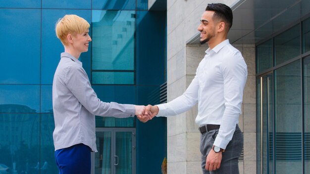 Two multiethnic colleagues Arabic man and caucasian woman stand outdoors talking shaking hands