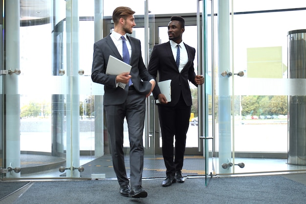 Two multinational young businessmen entering in office building with glass doors.