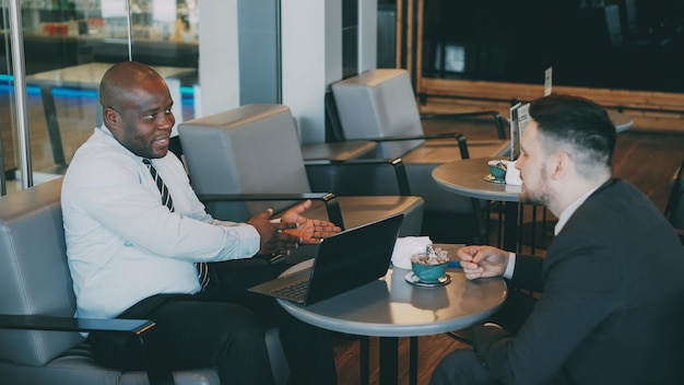 Two multiracial businessmen in formal clothes smiling gesturing and discussing their startup in spacious cafe during lunch break Advanced laptop is on their table