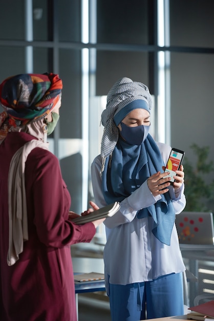 Two muslim businesswomen in hijabs using application on mobile phone during work at office