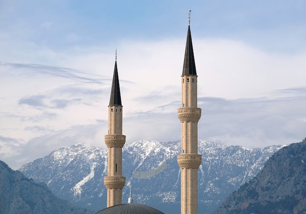 Two muslim minarets rise against impregnable snow-capped mountains overgrown at far with vegetation under serene blue sky