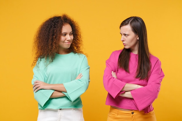 Two offended women friends european african girls in pink green clothes posing isolated on yellow background. people lifestyle concept. mock up copy space. holding hands crossed looking at each other