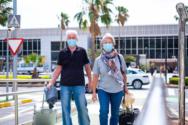 Two old seniors traveling together after quarantine and lockdown wearing medical and surgical mask to prevent covid-19 or anny type of new flu or virus - mature people with suitcase