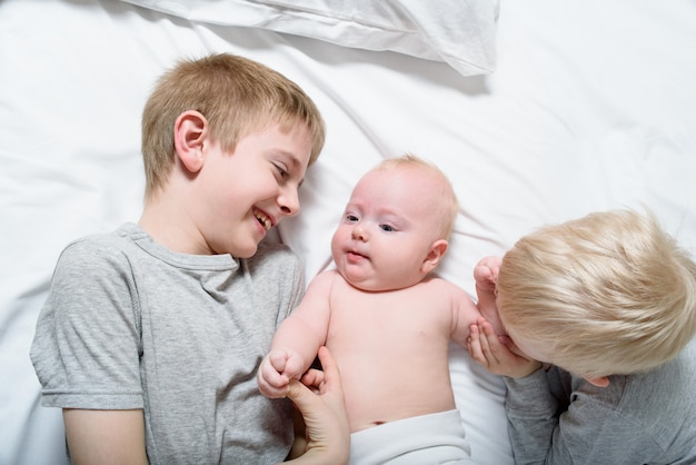 Two older brothers play and laugh with the youngest baby in a white bed. Happy childhood, big family