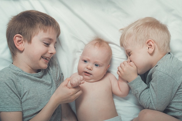 Due fratelli maggiori baciano e abbracciano teneramente il bambino più piccolo su un letto bianco. famiglia felice