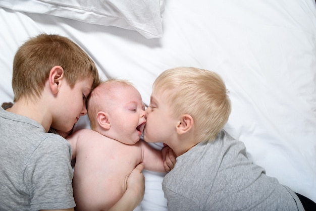 Two older brothers tenderly kiss and hug the younger child on a white bed.