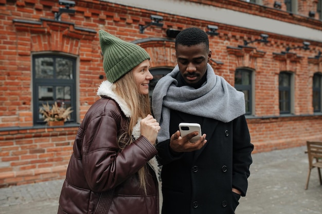 Two opposite sex interracial friends hanging out on the street, chatting, smiling. Both dressed warmly for late autumn. Man is staring at his phone.