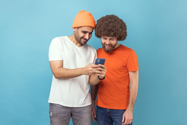 Двое оптимистичных мужчин стоят с помощью смартфона и читают позитивные новости в социальных сетях, улыбаясь