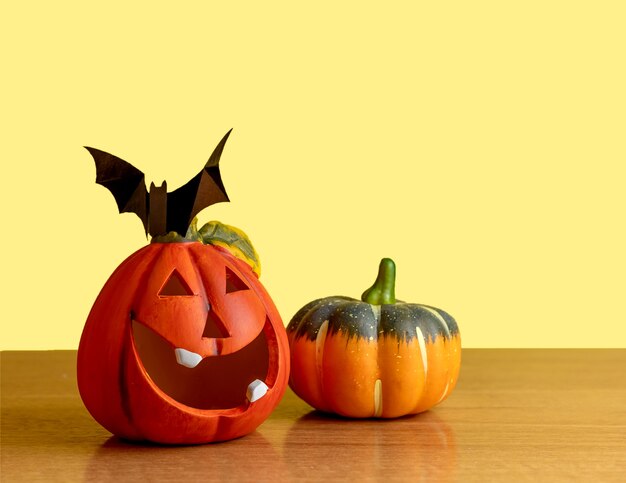 Two orange pumpkins stand on a table on a yellow background. A black bat sits on a pumpkin.