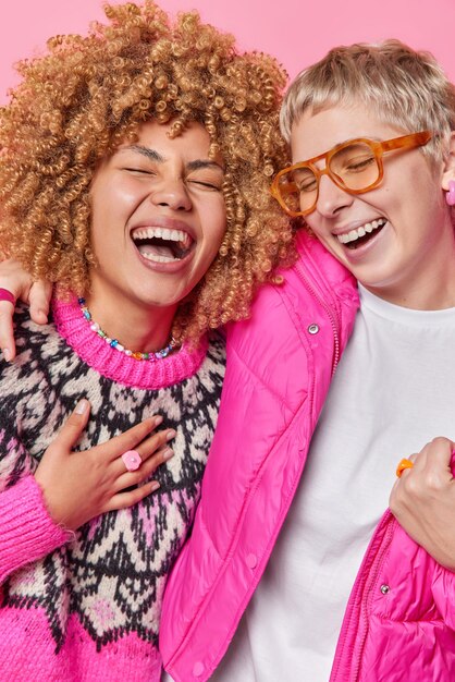 Two overjoyed young women have fun laugh happily express\
positive emotions and feelings embrace foolish around dressed in\
fashionable outfit isolated over pink background friends\
happiness