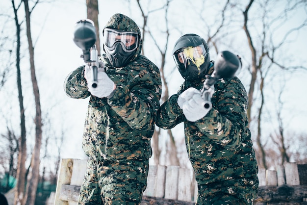 Two paintball players in splattered masks, team poses after winter battle. Extreme sport game, soldiers in special uniform, paintballing