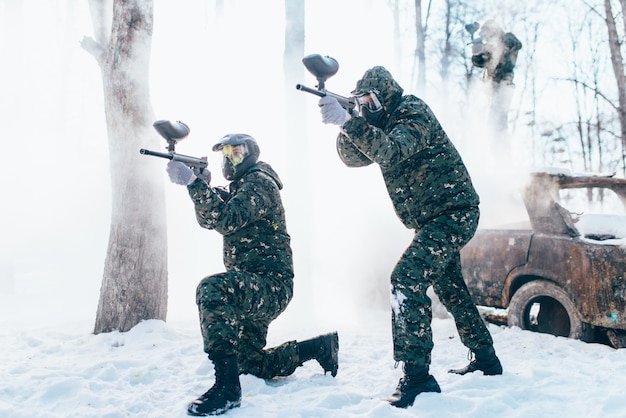 Two paintball players in uniform and masks shooting at the enemy, side view, winter forest battle. Extreme sport game