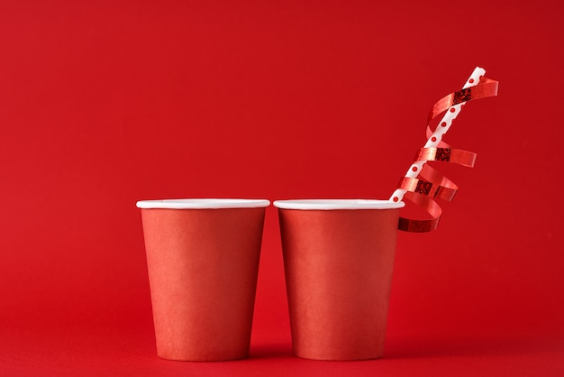 Two paper coffee cups with decorations and straw on red background.