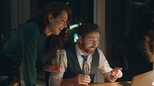 Two partners celebrating victory with champagne looking at laptop late night