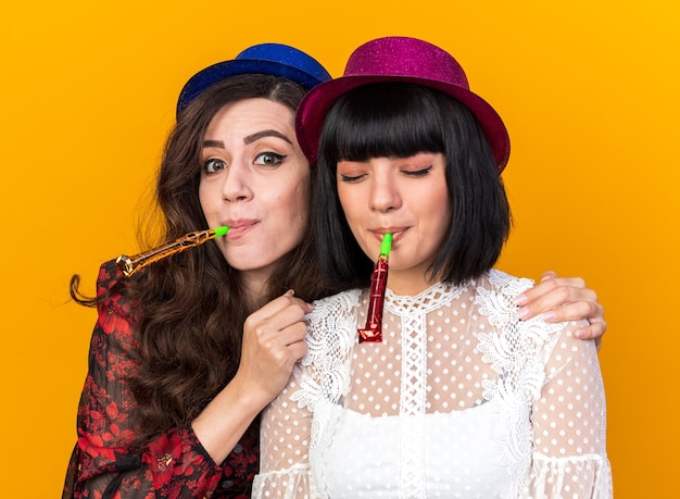 Two party girls wearing party hat one holding another one by shoulders  another with closed eyes both blowing party horn isolated on orange wall