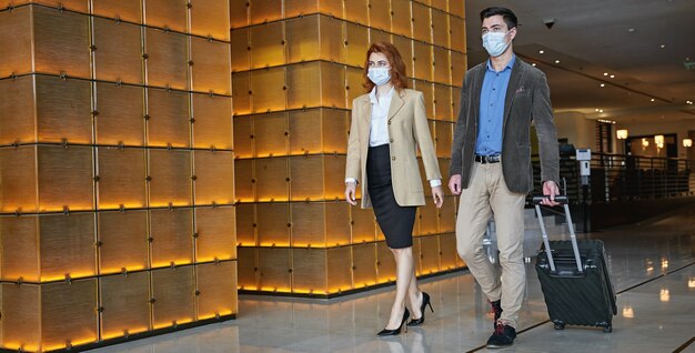 Два человека в медицинских масках идут по холлу отеля с чемоданом на колесиках. баннер веб-сайта