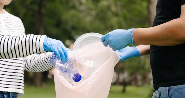 Два человека держат пластиковую бутылку с мусором в черном мешке в паркеОчистка экологии загрязнения и пластиковой концепции xA
