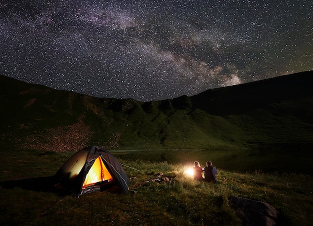 星と明るい天の川が散らばっている空の下で湖の近くの山でキャンプする夜の二人