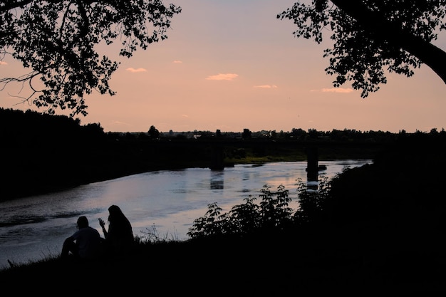 Two people sit by the river at sunset