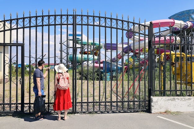 Two people stand at closed gates of abandoned water park and look through bars