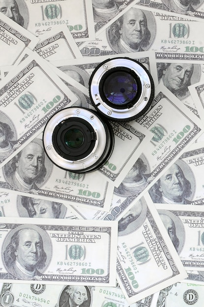 Two photographic lenses lie on the background of a lot of dollar bills. 