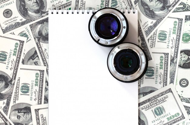 Two photographic lenses and white notebook lie on the background of a lot of dollar bills. Space for text