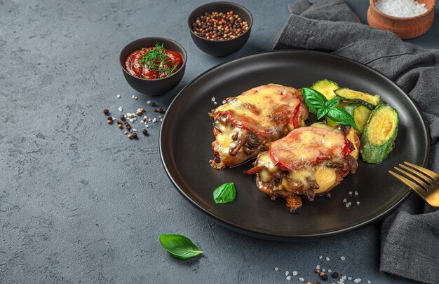 Two pieces of baked meat with mushrooms, tomato and cheese and a side dish of zucchini. in a black plate on a gray background. Side view, space for copying.